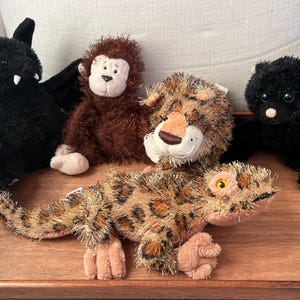 Puede incluir: Cinco peluches, incluyendo un murciélago negro, un mono marrón, un leopardo, un gato negro y un lagarto marrón y beige, están dispuestos sobre una superficie de madera.