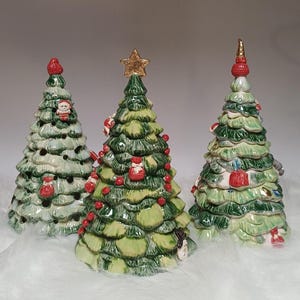 Könnte beinhalten: Drei weihnachtliche Tannenbaumfiguren aus Keramik in verschiedenen Grüntönen. Die Bäume sind mit kleinen roten Ornamenten, Geschenken und Figuren verziert. Ein Baum hat einen goldenen Stern als Spitze, die anderen haben rote Spitzen.