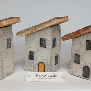 Op de afbeelding: Drie keramische huisjes met een witte craquelé glazuur, donkergrijze ramen en oranje of grijze boogdeuren. Elk bouwwerk heeft een schuin, getextureerd bruin dak. Een wit papieren label aan de voorzijde toont de tekst handmade with love.