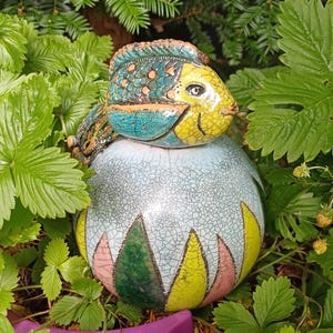 Peut inclure: Une sculpture en céramique composée d'une base ronde à glaçure craquelée bleu clair avec des motifs triangulaires alternés jaunes et roses à la base, surmontée d'une figurine de poisson stylisée aux tons turquoise, jaune et brun texturé.