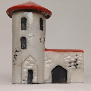 Op de afbeelding: Een keramisch miniatuurkasteel met een witte behuizing, een rood dak en zwarte, gewelfde deuren en ramen. De toren heeft drie ramen en de aangrenzende structuur heeft een gewelfde deur. Het kasteel heeft een rustieke, verouderde uitstraling.
