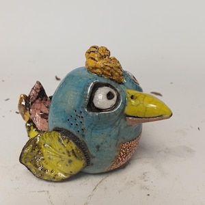 Peut inclure: Une figurine d'oiseau en céramique avec un vernis bleu craquelé, un bec jaune et une crête jaune texturée. L'oiseau présente un œil blanc avec une pupille noire, une aile jaune à motifs, une queue rose et une poitrine orange tachetée sur son corps rond.