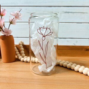 May include: A clear glass can-shaped drinking glass with a brown floral design. The glass contains white tissue paper. The glass sits on a wooden tray with a terracotta vase holding pink flowers and a string of wooden beads.