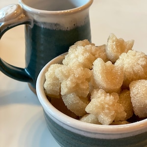 May include: A close-up of a bowl filled with light brown sugar cubes. The bowl is green and white with a white rim. The sugar cubes are in the shape of stars.