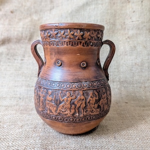 May include: A brown terracotta vase featuring two side handles and a wide rim. The upper section is decorated with a band of star-like motifs, while the lower body displays a detailed relief frieze of human figures and circular patterns.
