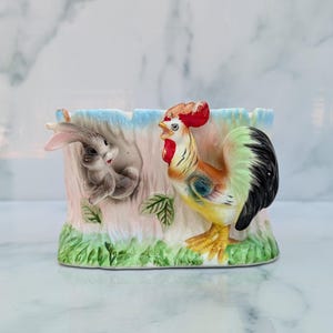 Könnte beinhalten: Ein Keramik-Pflanztopf in Form eines Baumstumpfes mit einem Hasen und einem Hahn, die herausgucken. Der Topf ist in Blautönen, Rosa, Grün und Braun bemalt.