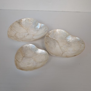 May include: Three white heart-shaped bowls made of mother of pearl. The bowls are different sizes and are arranged in a row.