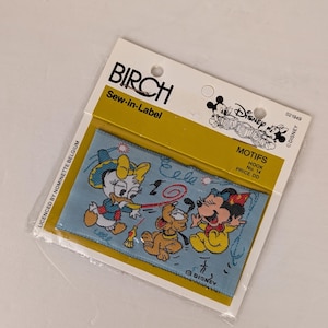 Op de afbeelding: Een blauw en geel naai-etiket met Disney-figuren Mickey Mouse, Minnie Mouse en Pluto. Het etiket heeft de tekst "Disney Babies" en "Motifs No. 14".