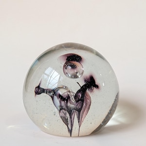 Può includere: Un fermacarte in vetro trasparente di forma sferica. All'interno, un design con forme astratte viola e nere. Il fermacarte ha una bolla sulla parte superiore ed è riempito di minuscole inclusioni scintillanti.