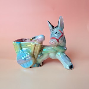 May include: A ceramic donkey planter with a cart, in iridescent blue, green, and pink. The donkey has a red bridle. The cart holds a small bird figure. A decorative item for home decor.
