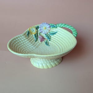 May include: A light yellow ceramic basket-shaped dish with a woven texture. The dish features a green handle and is adorned with colorful ceramic flowers and leaves. The basket sits on a pedestal base.