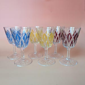 May include: A collection of six vintage crystal wine glasses. Each glass has a clear stem and base, with a coloured pattern in blue, yellow, and burgundy. The glasses are displayed on a light pink surface.