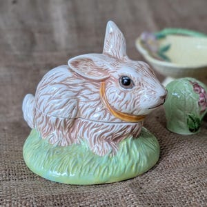 Könnte beinhalten: Keramik-Hasenfigur mit hellbraun-weißem Körper, sitzend auf einer grünen Grasbasis. Der Hase hat einen goldfarbenen Kragen und schwarze Augen. Weitere Keramikteile sind im Hintergrund zu sehen.