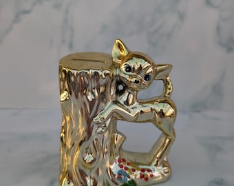 May include: A gold-coloured ceramic deer bank with a slot on top. The deer is posed next to a tree stump with colourful flowers at the base. The bank has a glossy finish and is a decorative item.