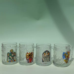 Pode incluir: Quatro canecas de vidro transparente com alças, cada uma com um design de vitral colorido diferente. Os desenhos incluem um corcunda, uma mulher de vestido vermelho, um anjo e um cavaleiro. As canecas têm um padrão texturizado.