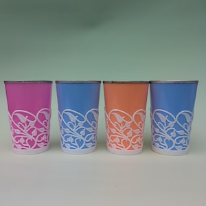 Op de afbeelding: Vier kleurrijke drinkglazen. Elk glas heeft een andere kleur: roze, blauw, oranje en blauw. Elk glas heeft een wit, decoratief bloemmotief rond de onderste helft. De glazen hebben een zilveren rand.
