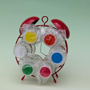 May include: A red metal spice rack in the shape of a clock, containing six glass spice jars with coloured lids. The lids are yellow, red, blue, green, pink, and white. The jars are wrapped in clear plastic.