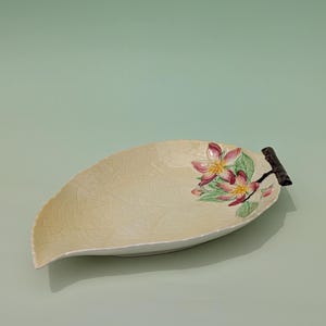 May include: Cream-colored leaf-shaped ceramic serving dish with a floral design. The dish features a textured surface and a dark brown handle. The floral design includes pink and yellow flowers with green leaves. A decorative piece for serving.