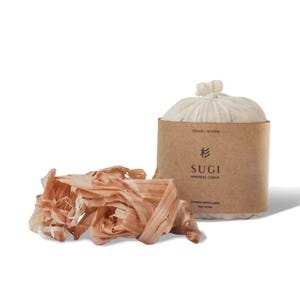 May include: A bundle of light brown wood shavings next to a cylindrical package wrapped in tan paper. The label reads Cedar-Myrrh, Sugi, Japanese Cedar, and Japanese Bath Flakes 28g / 0.99oz. The package is topped with a tied off-white fabric pouch.