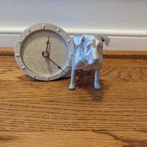 Op de afbeelding: Een zilverkleurige decoratieve klok met een hondenbeeldje. De klok heeft een ronde wijzerplaat met zwarte wijzers en een getextureerde rand. De hond staat naast de klok.