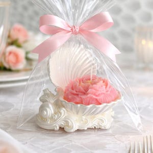 May include: A pink flower-shaped candle sits inside a white, shell-shaped holder. The entire item is encased in a clear plastic bag, which is tied at the top with a light pink satin ribbon bow, creating a decorative, gift-ready presentation.