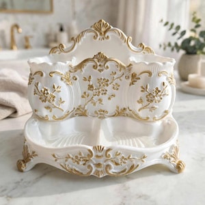 Könnte beinhalten: Ein weißer Kosmetik-Organizer mit goldenen floralen Reliefverzierungen. Das Objekt verfügt über zwei zylindrische Fächer oben und zwei flache, muschelförmige Schalen an der Basis, die auf vier verzierten, geschwungenen Füßen stehen.