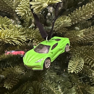Puede incluir: Adorno navideño de coche de juguete verde brillante con ruedas plateadas y una cinta negra para colgar. El adorno está suspendido sobre un fondo de ramas verdes de árbol de Navidad, creando una escena festiva.