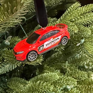 May include: A red Honda Civic Type R car ornament with the text "HONDA R" and the number "3" on the side. The car has a black ribbon for hanging and is displayed on a Christmas tree.