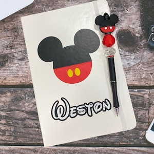 Puede incluir: Una libreta blanca con un diseño de Mickey Mouse y el nombre "Weston" en una fuente de Disney. La libreta tiene un bolígrafo negro con un dije de cabeza de Mickey Mouse.