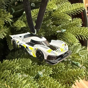 Op de afbeelding: Een wit en zwart raceauto-ornament met gele accenten hangt aan een zwart fluwelen lint. Het ornament toont het woord "Peugeot" en hangt voor een groene kerstboom.