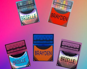 Op de afbeelding: Vijf gepersonaliseerde kleurpotloodoosjes in verschillende kleuren, elk met een naam. De doosjes zijn open en tonen de kleurpotloden binnenin. De namen "Brielle" en "Brayden" staan op de voorkant van de doosjes, met een holografisch ontwerp.