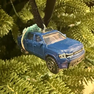 May include: A blue toy pickup truck Christmas ornament. The truck has black wheels and a black roof. The ornament is hanging from a black ribbon and is surrounded by green pine needles.