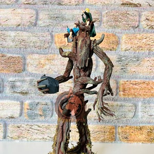 May include: A brown tree-like figure with a face and long branches, holding a dark gray rock in one hand. Two small figures are sitting on the tree's head. The figure is standing on a round base with green moss.