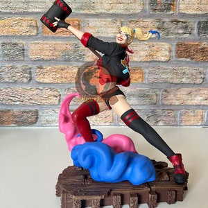 Pode incluir: Uma estatueta de uma personagem com marias-chiquinhas loiras vestindo uma jaqueta preta, top vermelho e shorts pretos, segurando um grande martelo preto e vermelho acima da cabeça. A figura está sobre uma base de fumaça azul e rosa esculpida.