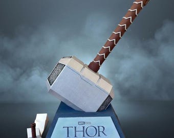 Könnte beinhalten: Ein großer silberfarbener Hammer mit einem braunen Griff mit weißem Zickzackmuster, montiert auf einem blauen Sockel mit einer Plakette mit der Aufschrift Marvel Studios THOR MJOLNIR. Eine Miniaturversion des Hammers steht links vom Sockel.