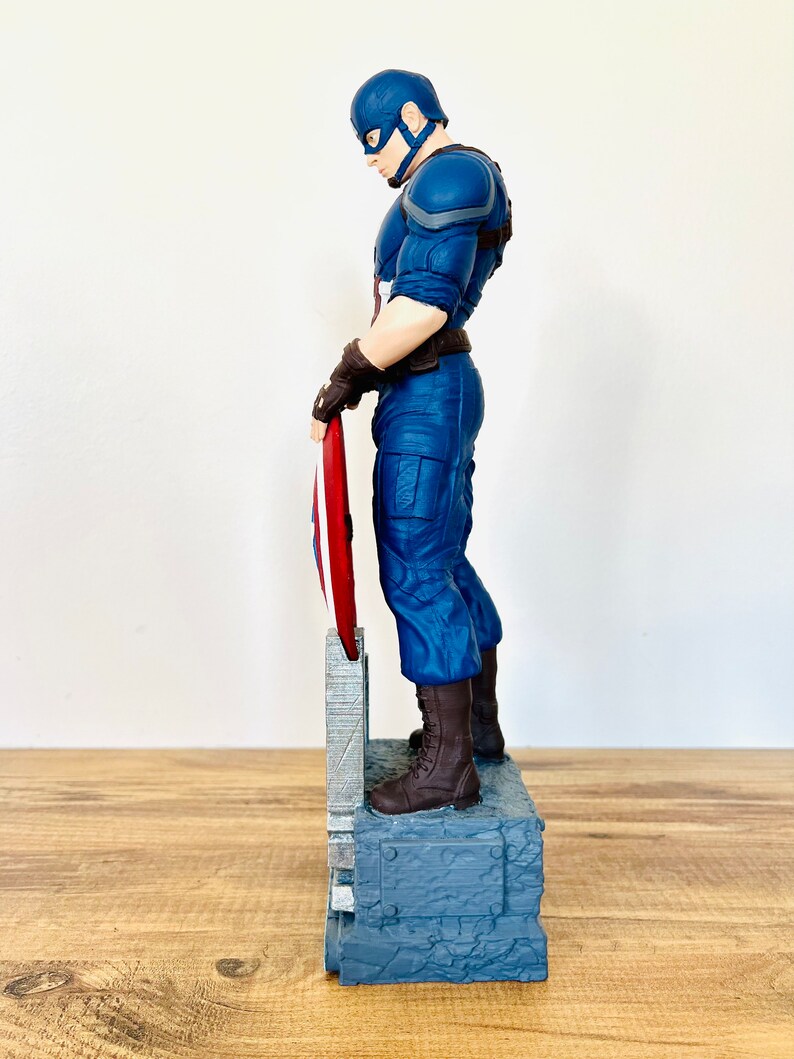 May include: A Captain America figurine in a blue and red costume, holding his shield. The figurine is standing on a gray base with a textured design. The figure is approximately 12 inches tall.