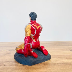 Pu&ograve; includere: Una action figure rossa e dorata di un supereroe in posizione inginocchiata. La figura &egrave; su una base nera.