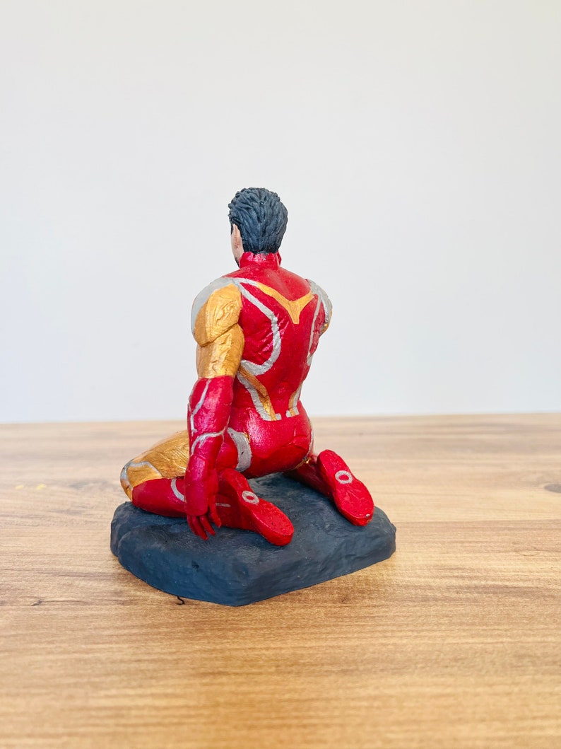 Pu&ograve; includere: Una action figure rossa e dorata di un supereroe in posizione inginocchiata. La figura &egrave; su una base nera.