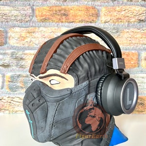 May include: A black and brown sculpted head wearing a black and brown headset with a black and silver earcup. The head is wearing a black mask with a blue and white design. The background is a brick wall.