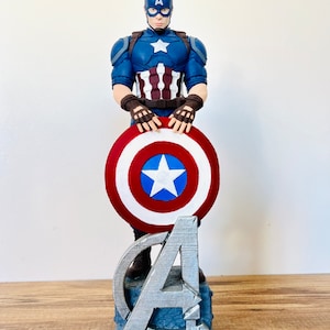 May include: A Captain America figurine, featuring a blue helmet and suit with a white star. The figure holds a red, white, and blue shield. The base has the Avengers logo and the word "LEGEND".
