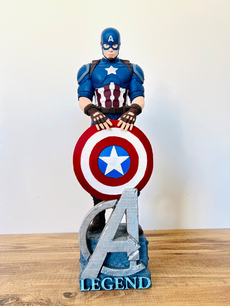 May include: A Captain America figurine, featuring a blue helmet and suit with a white star. The figure holds a red, white, and blue shield. The base has the Avengers logo and the word "LEGEND".