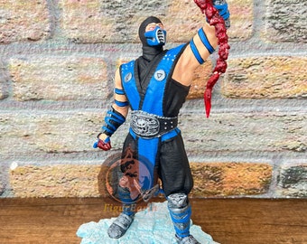 May include: A Sub-Zero figurine standing on a light blue cracked ice base. The character wears a black and blue ninja outfit with a silver belt buckle and holds a severed head with a spinal column attached, featuring red blood details.