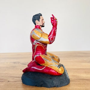 Pu&ograve; includere: Una action figure di Iron Man inginocchiato con la mano destra alzata, che tiene il Guanto dell'Infinito con tutte e sei le Gemme dell'Infinito. La figura &egrave; rossa e dorata e si trova su una base nera.