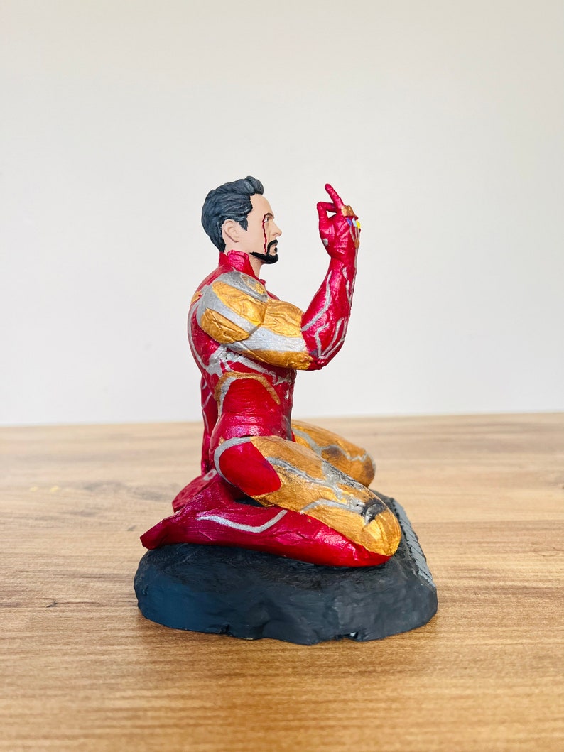 Pu&ograve; includere: Una action figure di Iron Man inginocchiato con la mano destra alzata, che tiene il Guanto dell'Infinito con tutte e sei le Gemme dell'Infinito. La figura &egrave; rossa e dorata e si trova su una base nera.