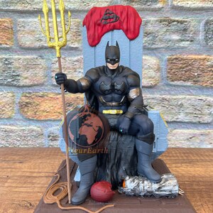 May include: A Batman figurine seated on a gray throne with a red cloth draped over the top. The figure wears a black suit with gold accents, holds a tall gold trident, and rests one foot on a silver metallic object. A small red sphere sits nearby.