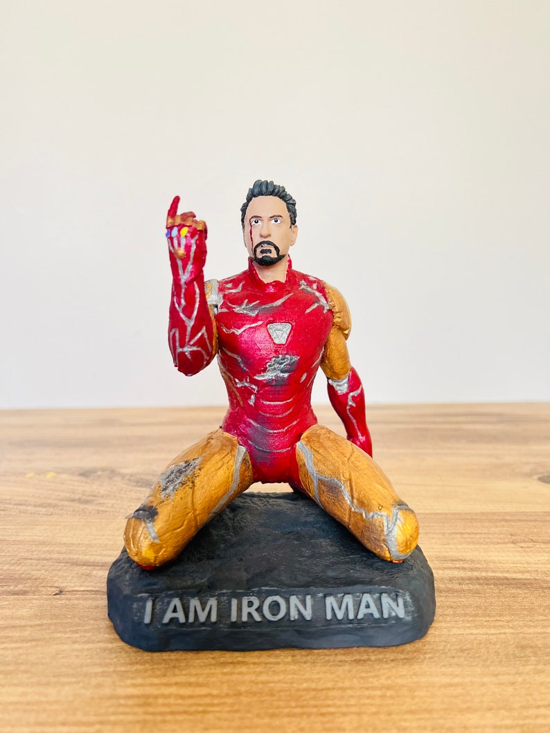 Pu&ograve; includere: Una action figure di Iron Man rossa e dorata inginocchiata con il Guanto dell'Infinito in mano. La figura &egrave; danneggiata e ha una base nera con le parole "I AM IRON MAN" scritte su di essa.