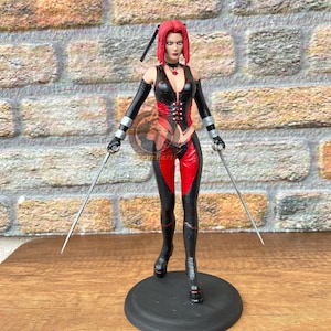 Peut inclure: Une figurine articulée debout aux cheveux rouges courts, vêtue d'un gilet sans manches noir et rouge avec un legging assorti. La figurine porte de longs gants noirs, un collier à pendentif rouge et tient une fine lame argentée dans chaque main.