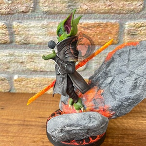 May include: A green and black goblin figurine with large ears, holding a black orb and standing on a rock base with orange flames. The figurine is wearing a black cloak and armor.