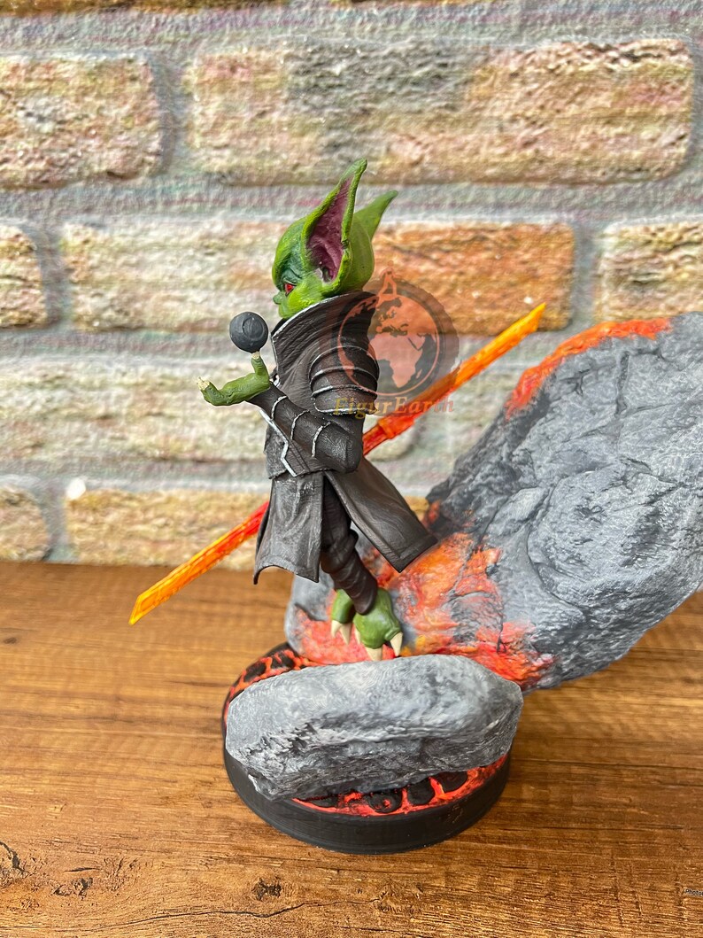 May include: A green and black goblin figurine with large ears, holding a black orb and standing on a rock base with orange flames. The figurine is wearing a black cloak and armor.