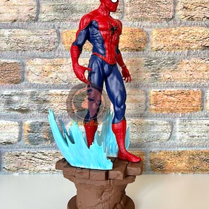 May include: A red and blue Spider-Man action figure stands on a brown stone base with blue and white web-like effects. The figure is posed with one arm raised and the other at its side.